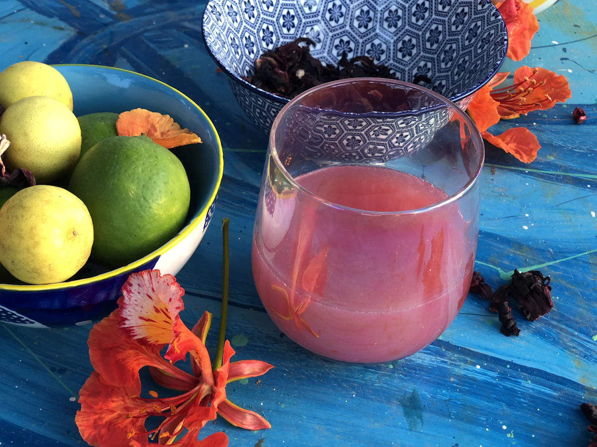 A glass jug of deep pink sorrel next to limes and dried hibiscus